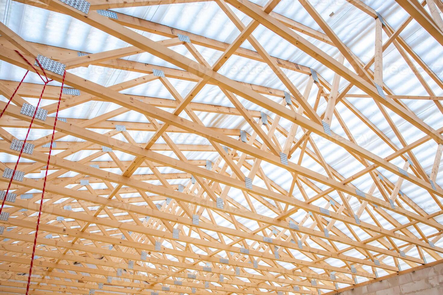 Wooden truss roof under construction with light through translucent panels, metal connectors at joints, and a red cable hanging on the left side. photo