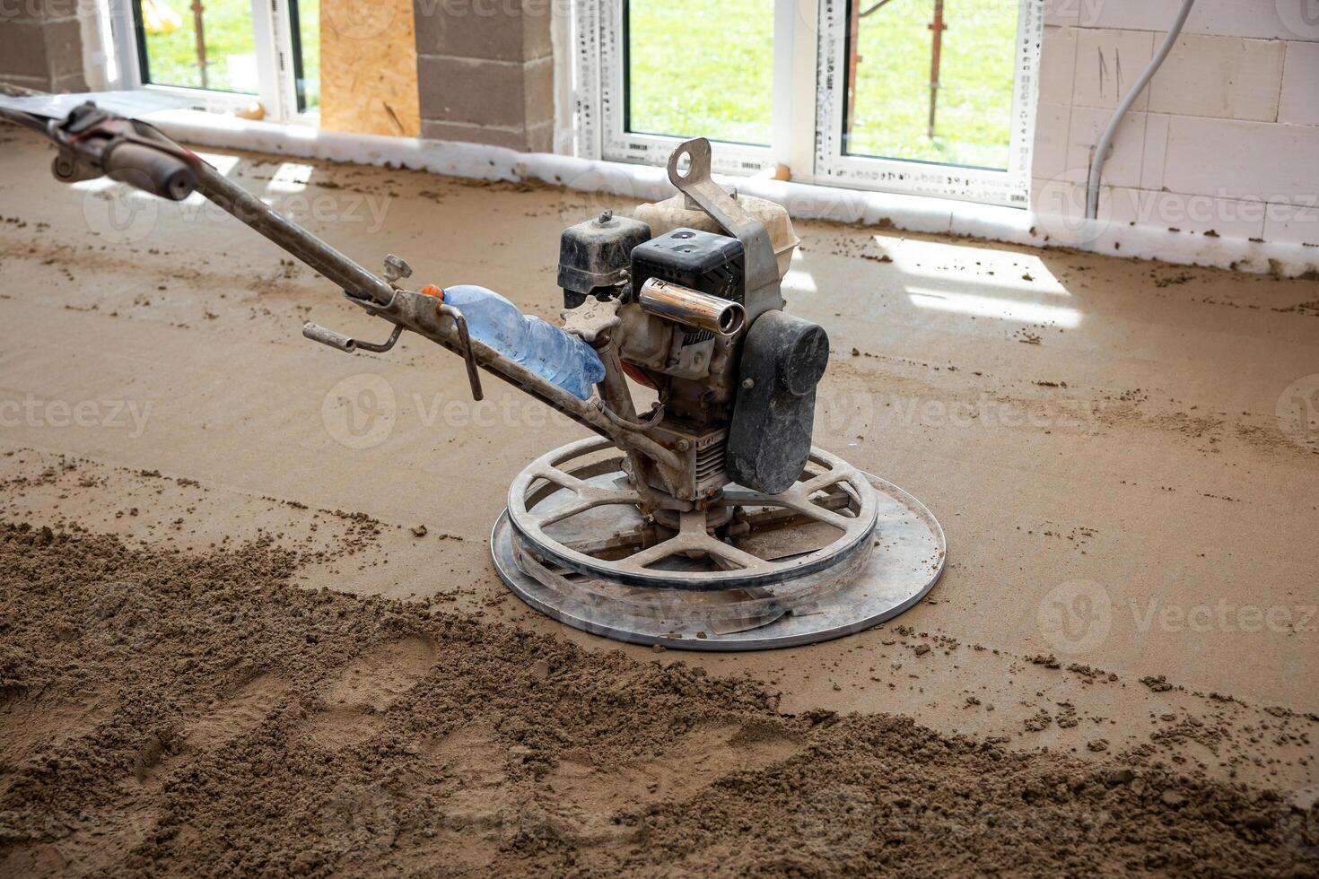 Construction site interior with a power trowel machine smoothing concrete over underfloor heating pipes. Unfinished walls and large windows are visible. photo