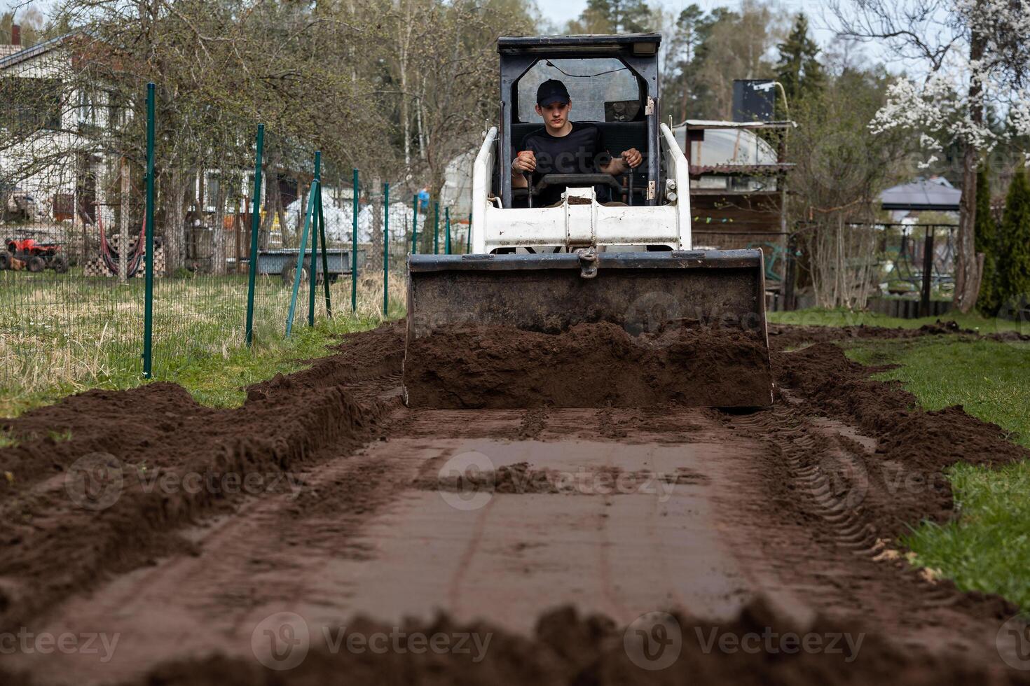 A man uses a white bulldozer to level soil on a narrow pathway bordered by grass, with a wire fence, houses, and trees visible in the distance. photo
