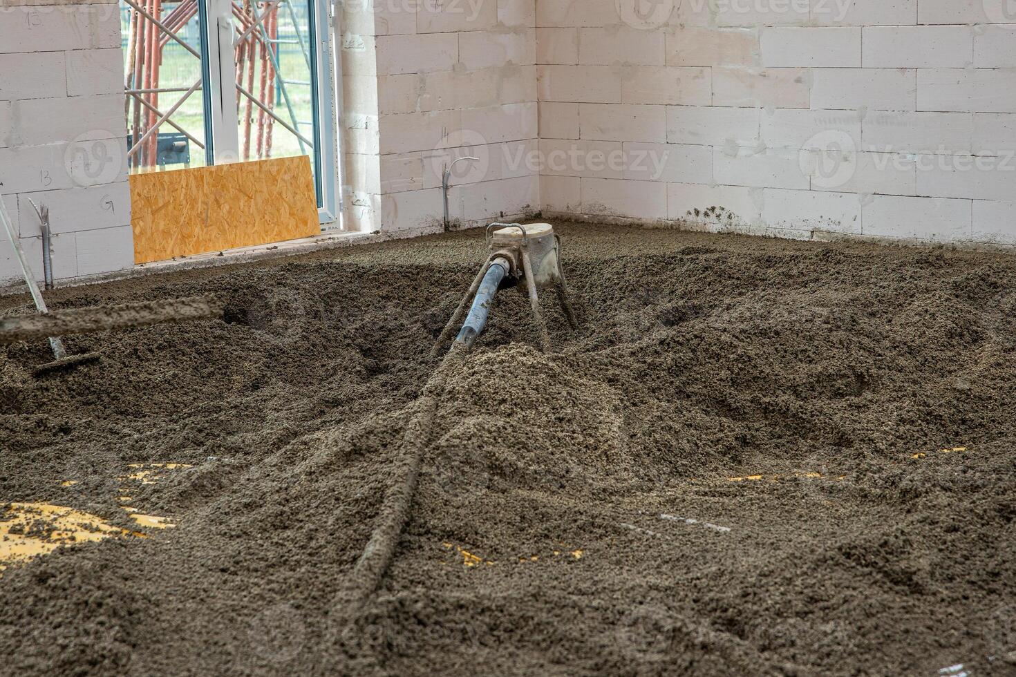 White brick walls and an unfinished floor being leveled with concrete. A hose distributes material, with light entering through a window opening. photo