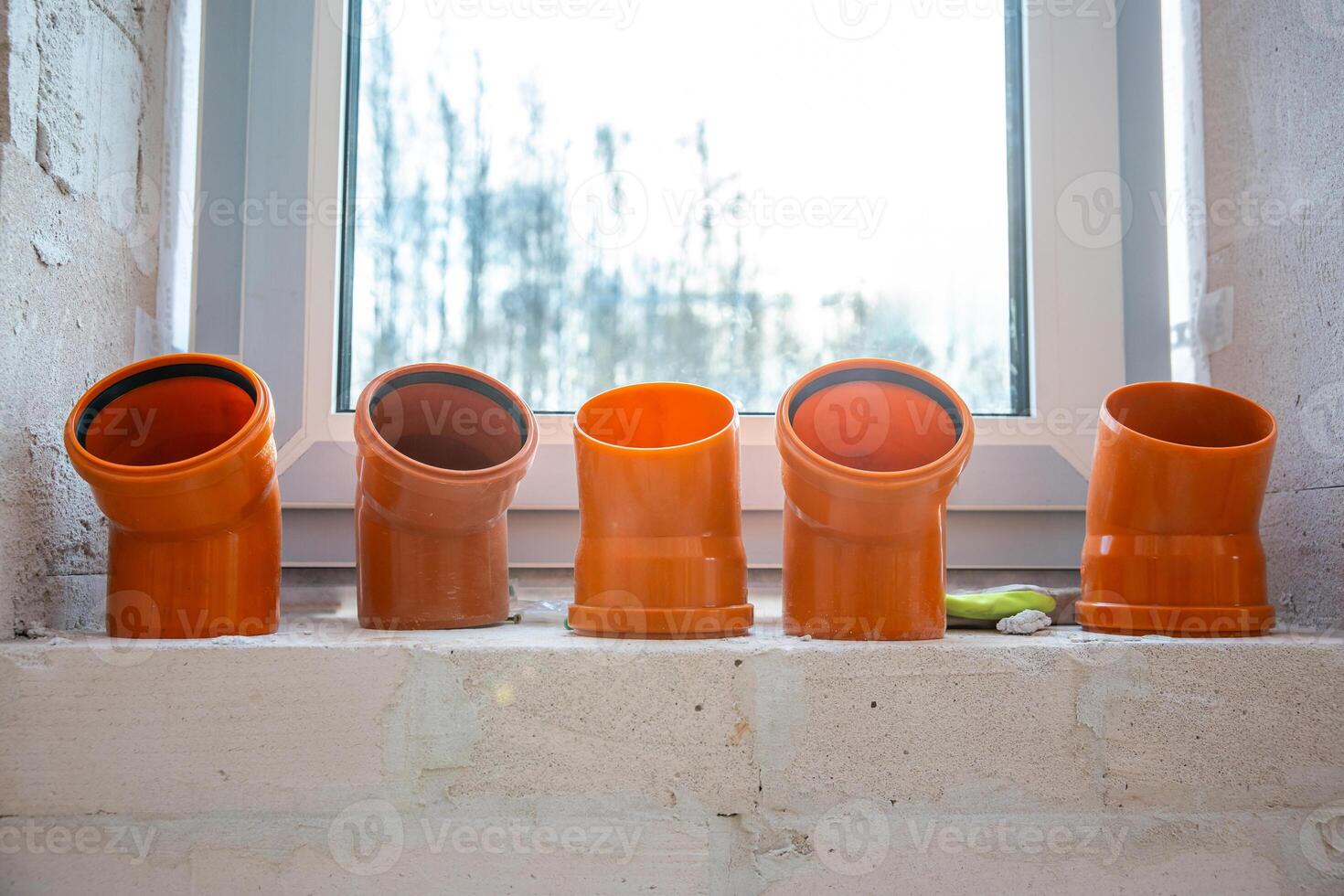 Five orange PVC pipe fittings sit in a row on a light concrete windowsill. A white framed window reveals blurred leafless trees outside in winter. photo