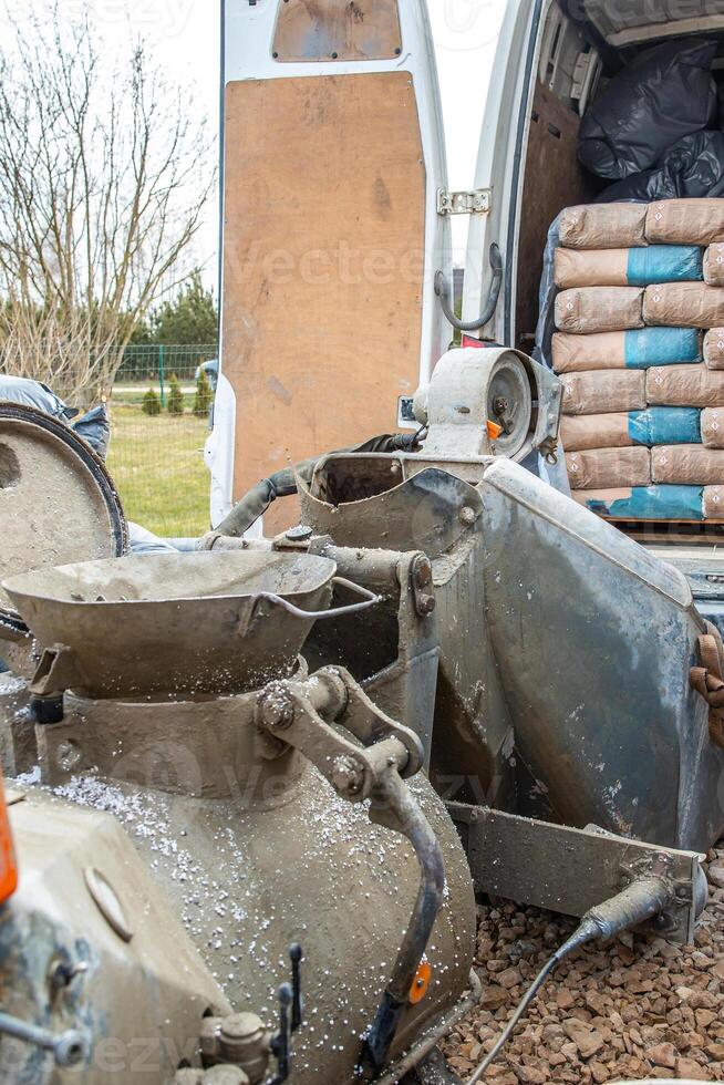 A construction site featuring a worn cement mixer, a van loaded with cement bags, gravel covered ground, sparse trees, and a distant fence. photo