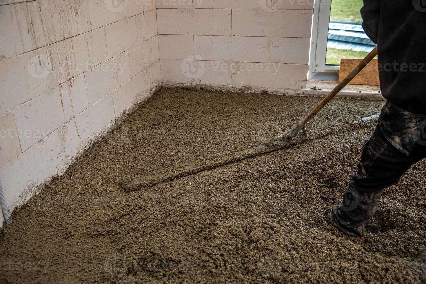 A construction worker smooths fresh concrete inside a building with white concrete block walls, an open window, and visible signs of ongoing work. photo