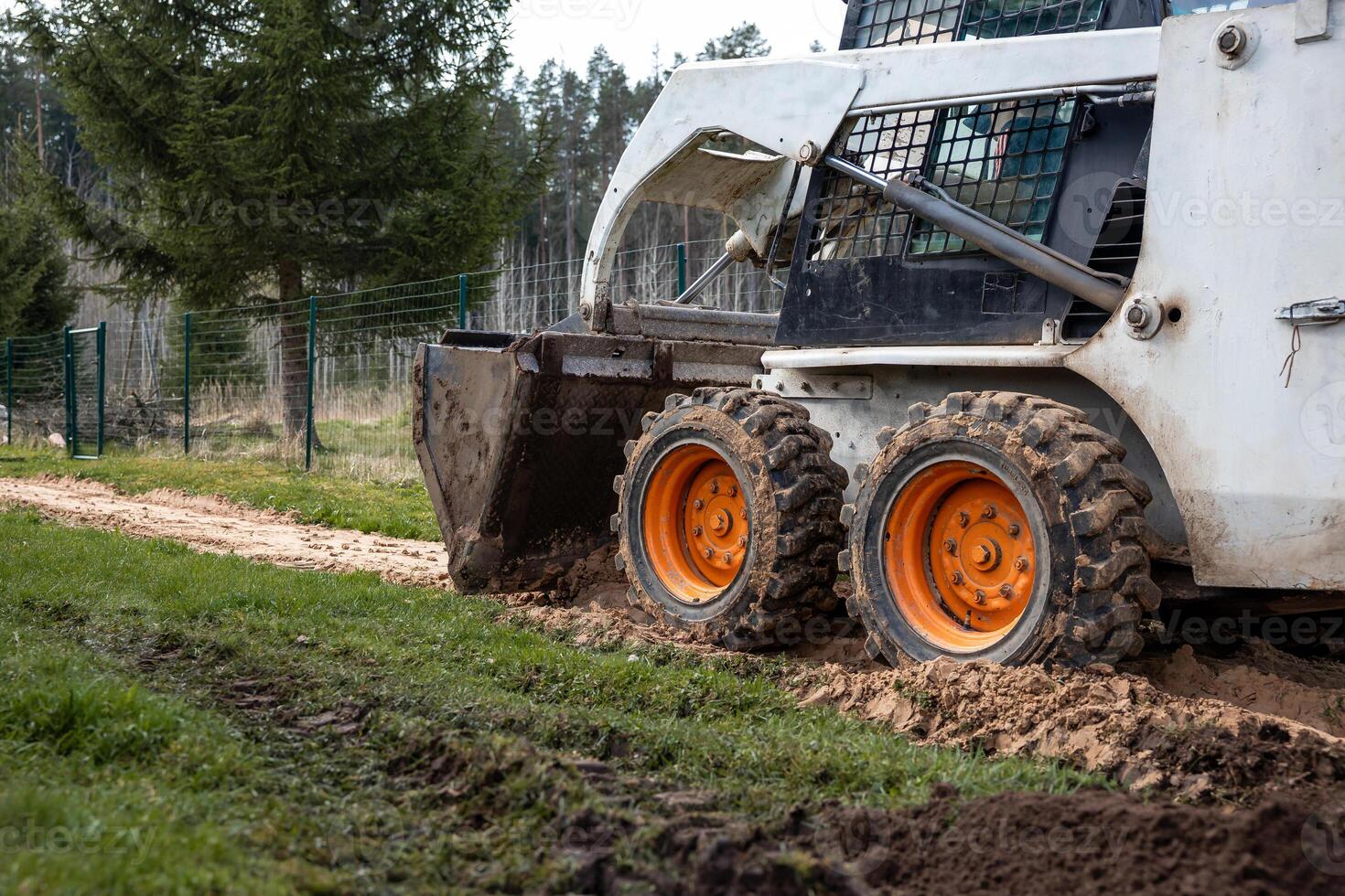 A skid steer loader with orange wheels works on a dirt path, leaving tire tracks. The bucket is lowered, with a wire fence and evergreen trees nearby. photo