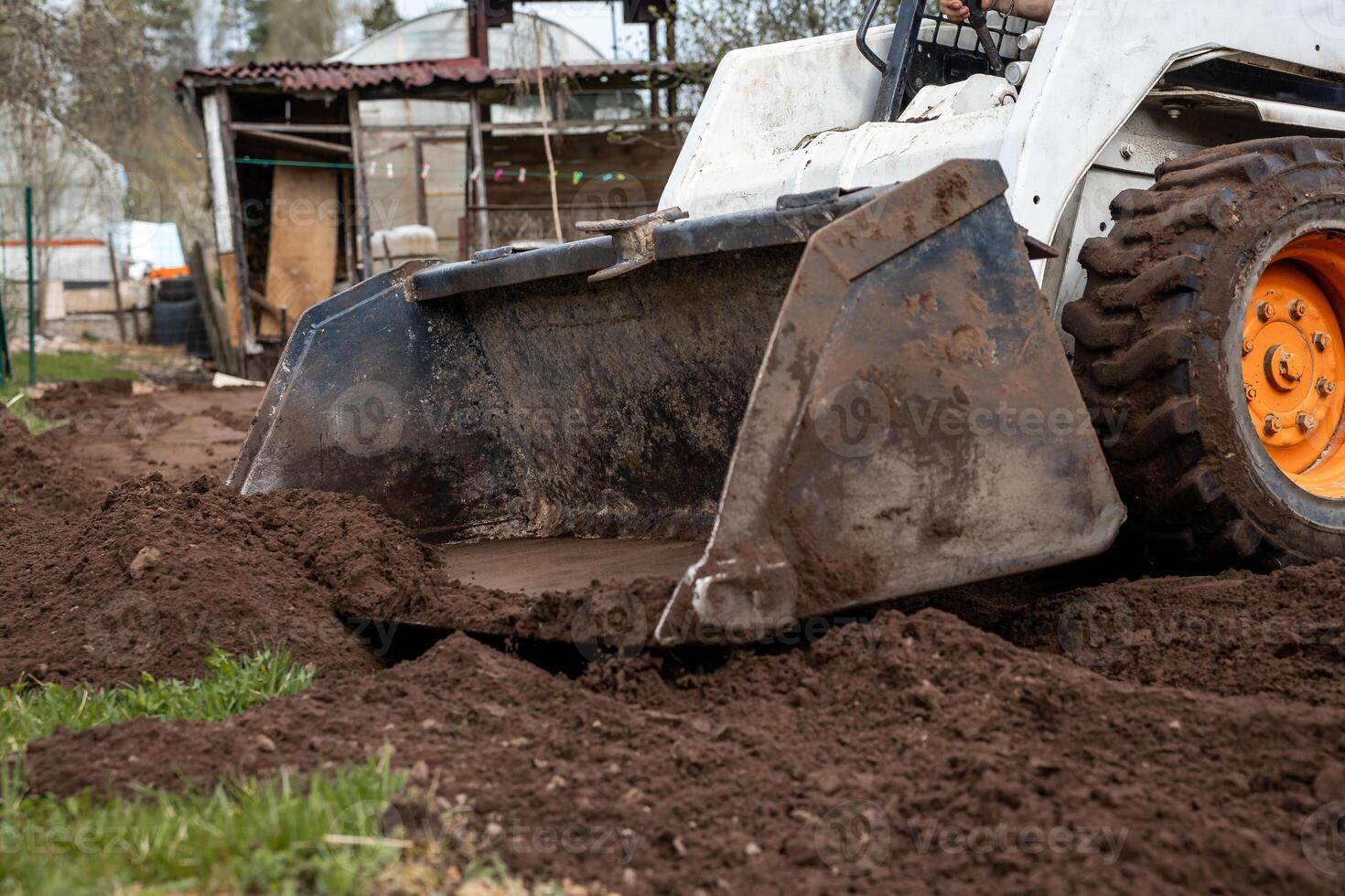 A small construction vehicle with an orange wheel and front loader moves soil near a rustic structure with a corrugated metal roof and wooden panels. photo
