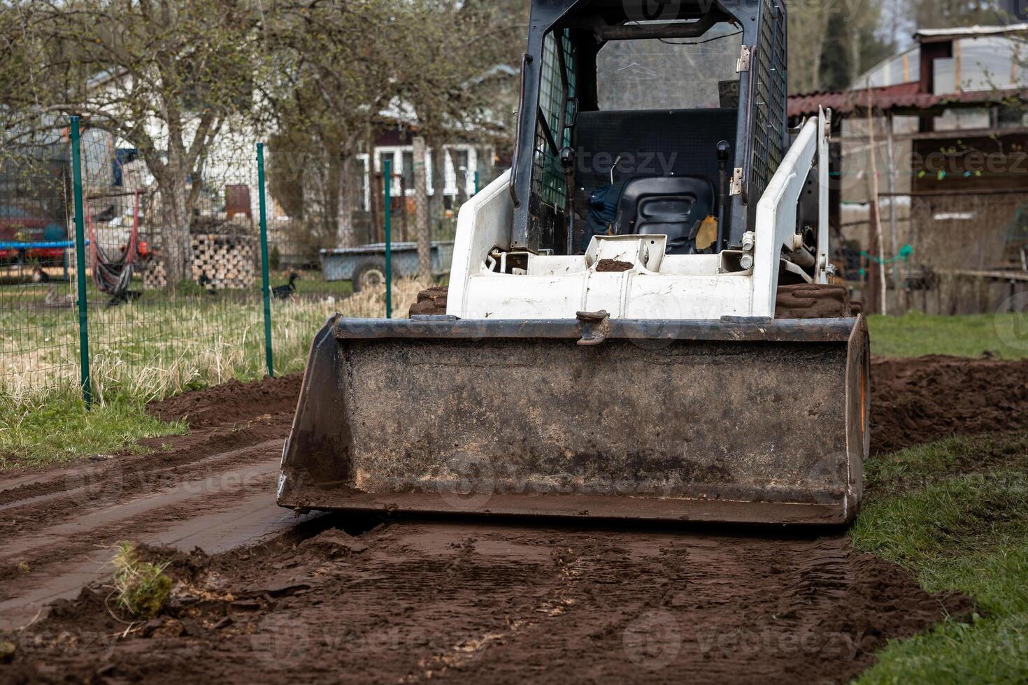 A white skid steer loader works on leveling soil on a dirt path, surrounded by fenced areas, small structures, and trees in a rural setting. photo