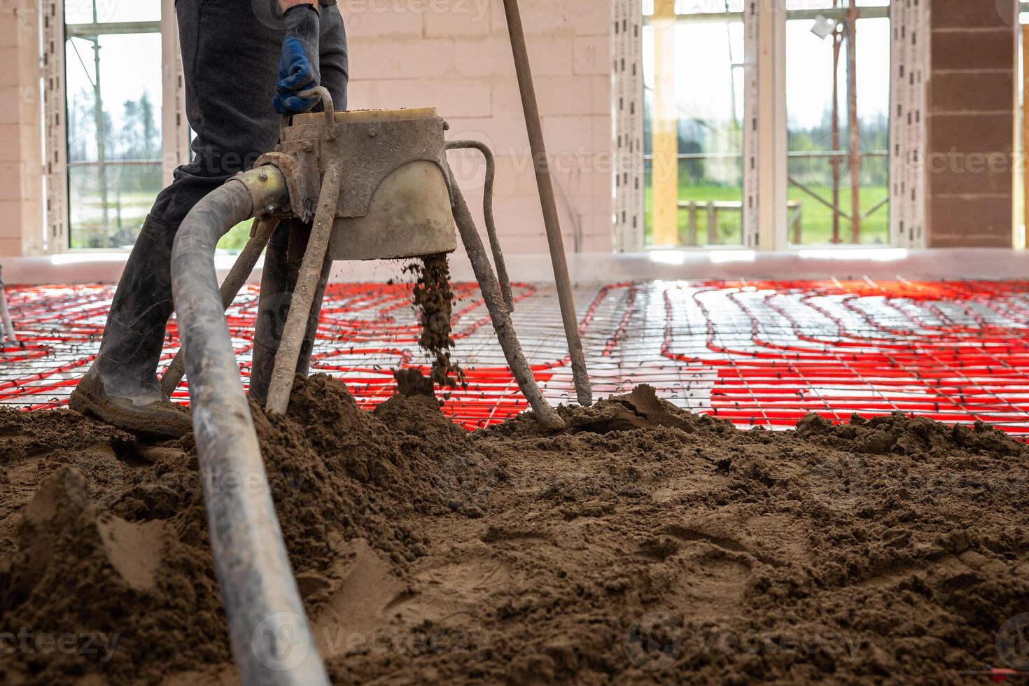 An indoor construction site shows underfloor heating pipes in a grid pattern being covered with Estrich concrete. A worker operates pouring equipment. photo