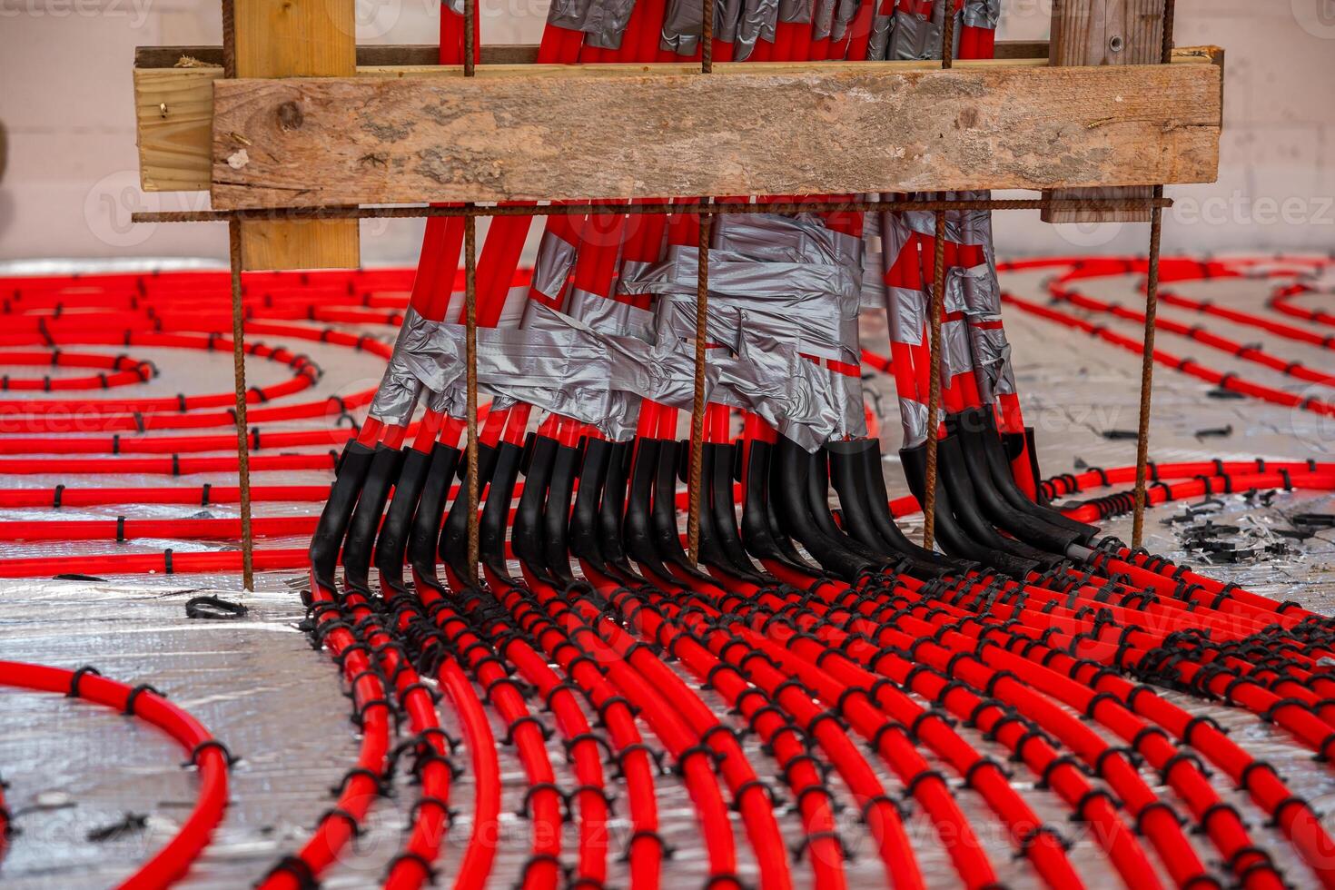 Bright red underfloor heating pipes arranged in a coiled pattern on a reflective surface, secured with duct tape and supported by a wooden frame. photo