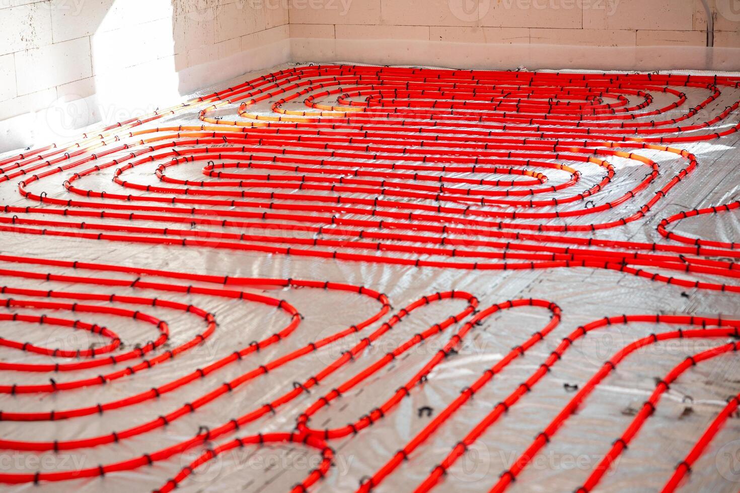 Bright red underfloor heating pipes are arranged in a looping pattern on a reflective surface, secured with clips, in a white block walled construction site. photo
