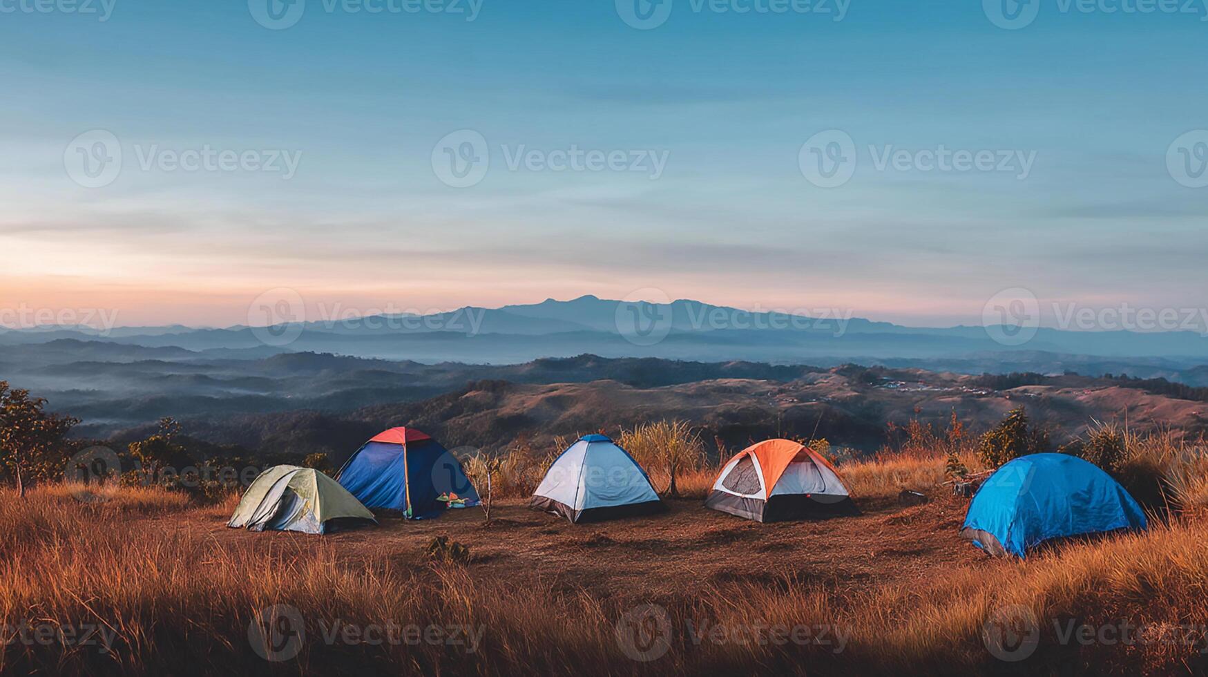 Multiple camping tents are arranged in a scenic mountainous area, surrounded by tall trees and wild grass. photo
