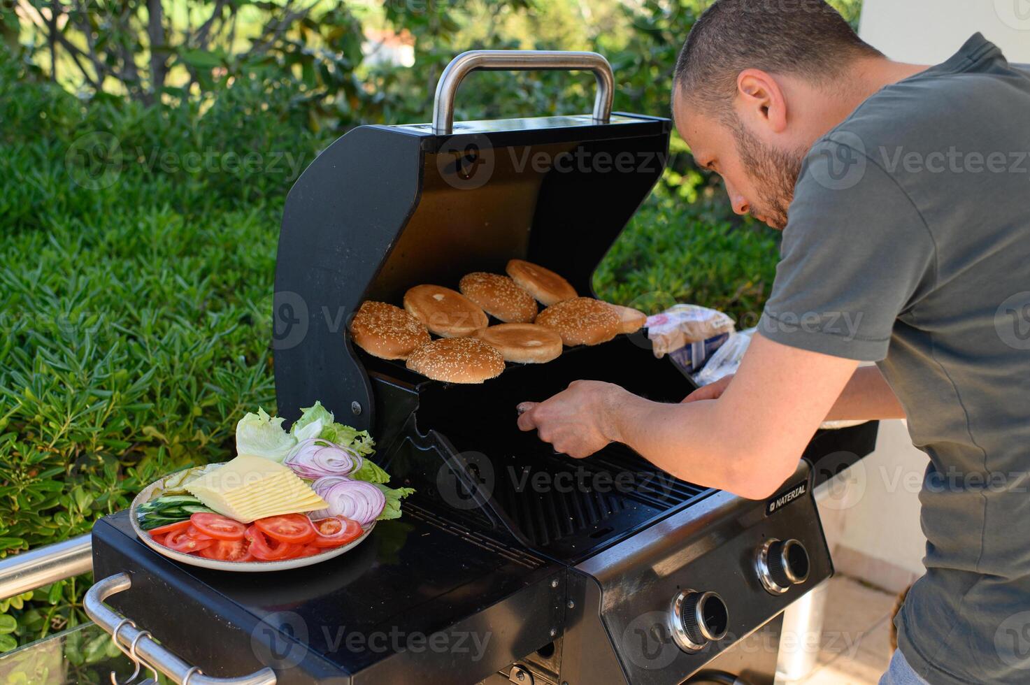 Grilling in the open air a man cooks beef patties and toasts buns next to a plate of crisp vegetables and cheese slices, ready for a summer feast. photo