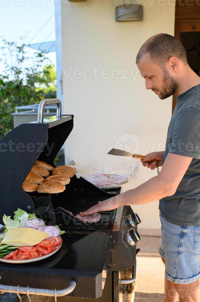 A man grills burger patties over an open flame, with sesame seed buns toasting nearby and a plate of fresh vegetables and cheese ready for burger assembly. photo