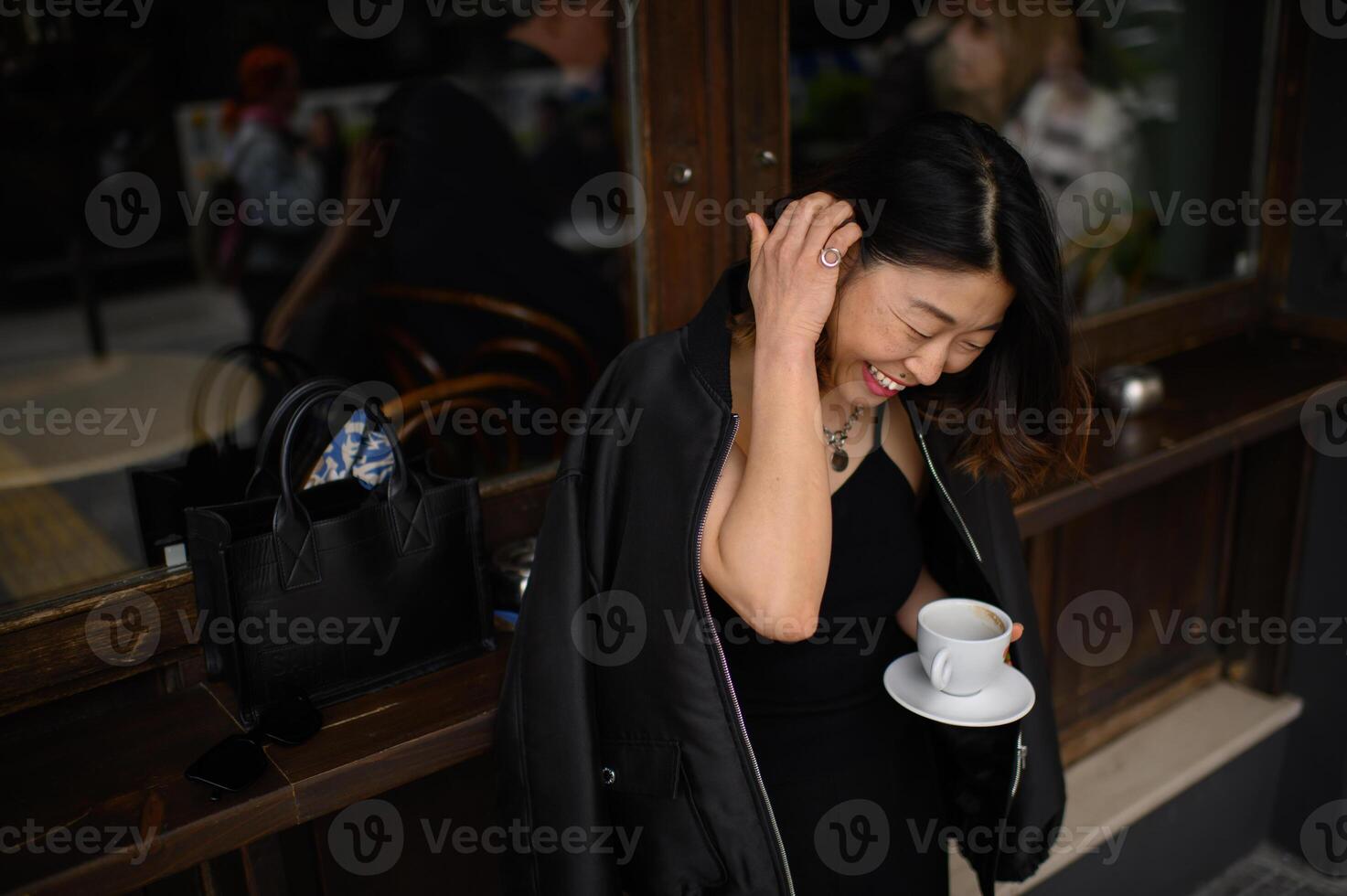 A cheerful Asian girl smiles widely as she stands outside a cafe in the summer. With a cup of coffee on a saucer in hand, her expression exudes carefree joy. photo