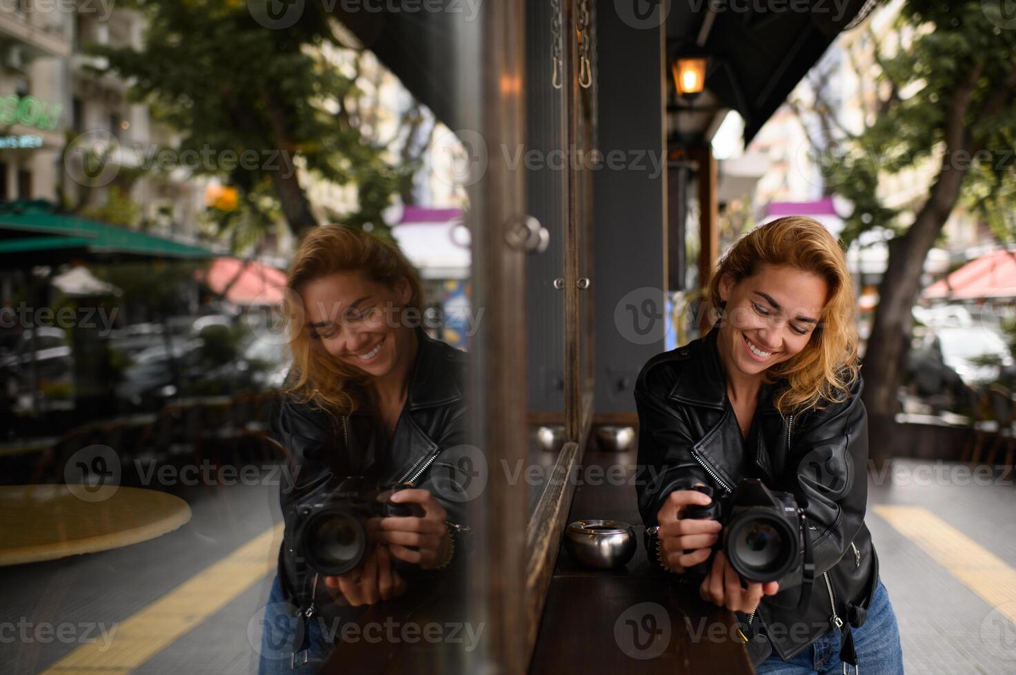Standing near a cafe, a woman holds her camera while her reflection appears in the glass window. The soft light and double image add depth to the urban atmosphere. photo