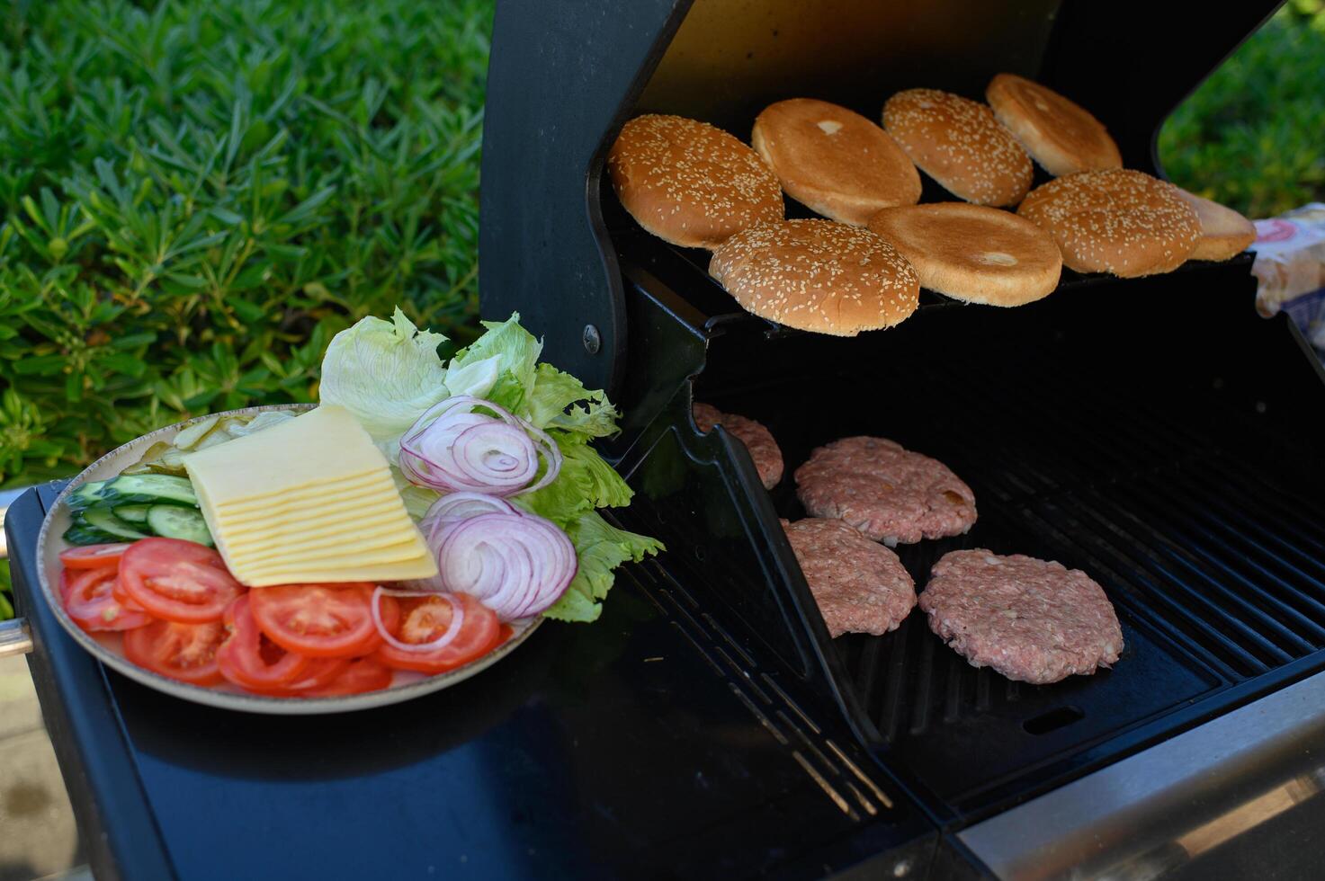 Juicy burger patties and golden buns are grilling outdoors, while a plate of fresh toppings sits ready for burger assembly on a warm summer day photo