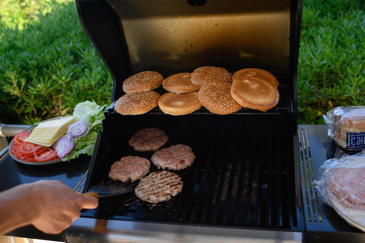 Hands flip sizzling burger patties on an outdoor grill, with buns toasting nearby and a plate of fresh vegetables and cheese ready for burger assembly photo