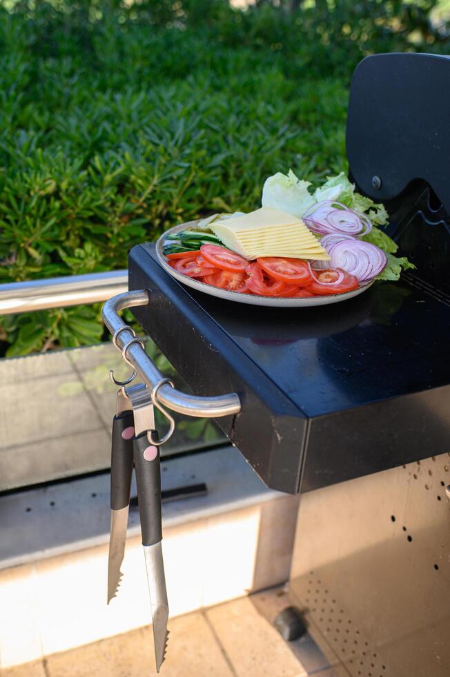 Colorful plates of crisp lettuce, tomatoes, onions, pickles, and cheese slices sit on the grill beside sizzling burger patties. photo