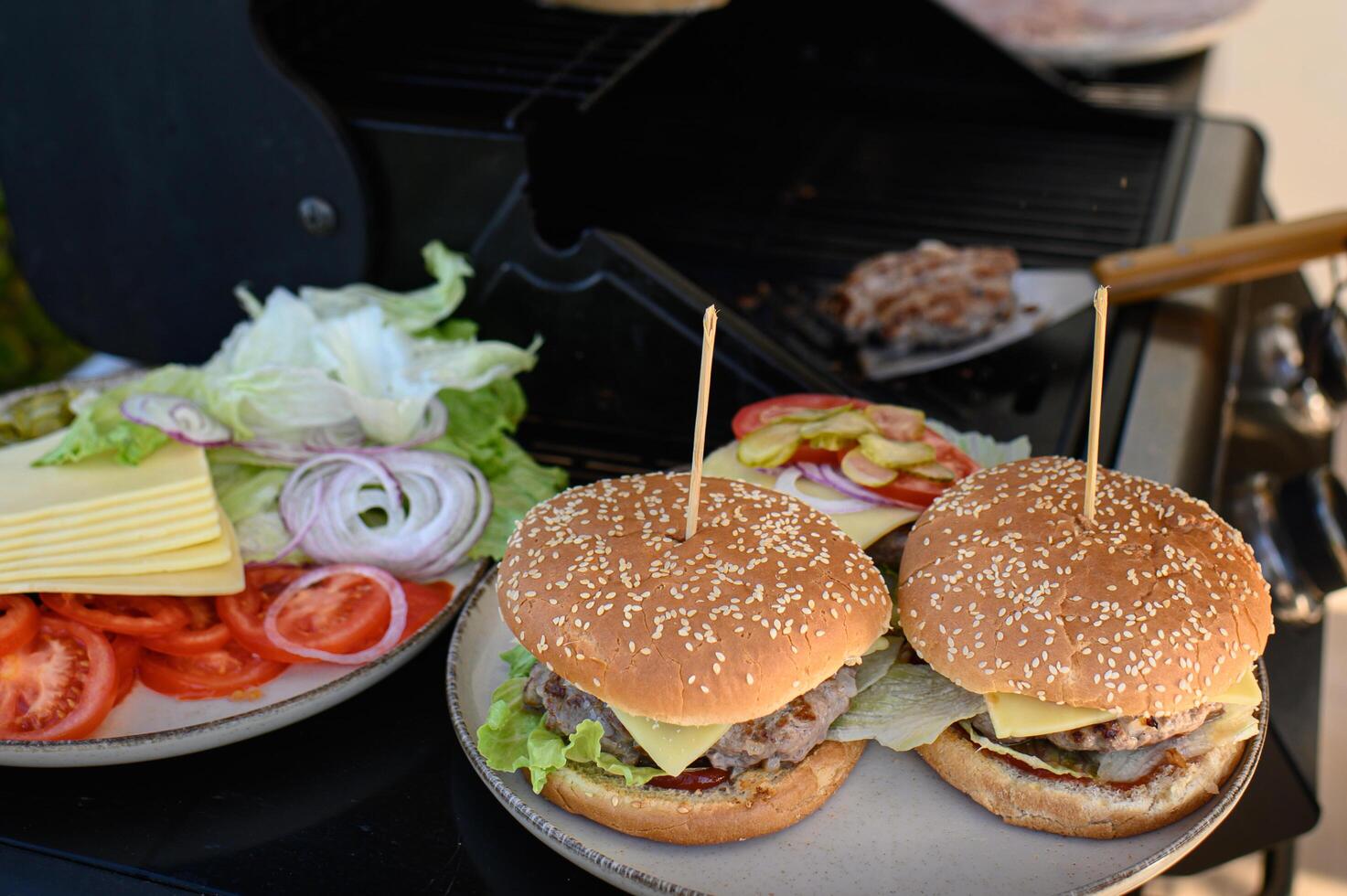 Outdoor burger making by the grill, with patties cooking, buns toasting, and burgers being finished with crisp veggies and cheese, each pierced by a skewer. photo