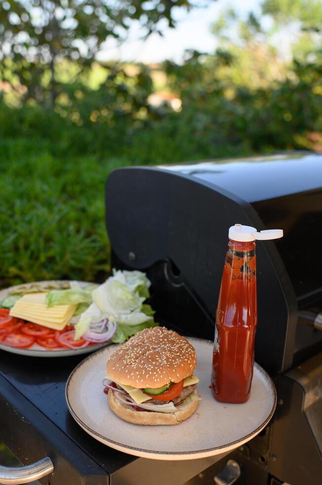 A freshly assembled burger stands next to a bottle of ketchup on the grill, with buns still toasting in the background. photo