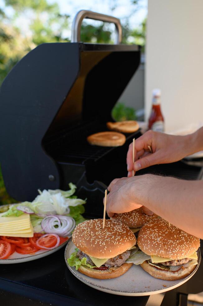 Hands assemble burgers next to a hot grill where patties are sizzling and buns are toasting, with a bowl of fresh vegetables and cheese nearby. photo