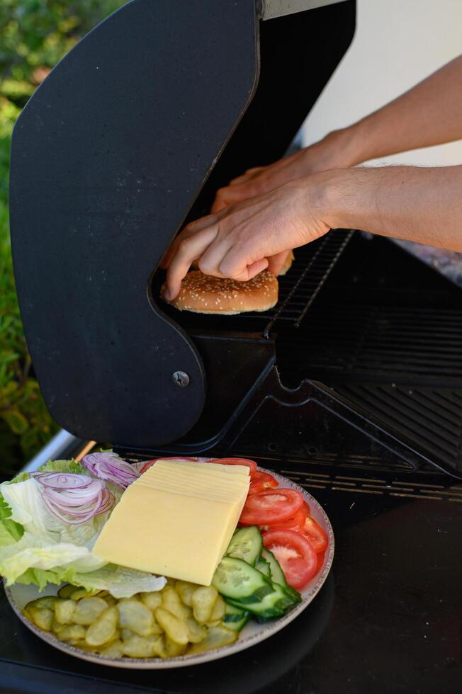 Hands place burger buns on a hot grill next to sizzling patties, with a plate of fresh vegetables and cheese ready for assembling burgers. photo