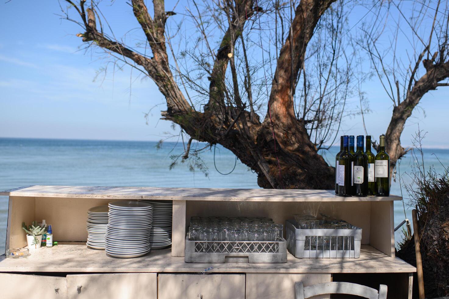 A charming bar in a Greek taverna, decorated with wine bottles and stacked plates, set under a tree with a stunning sea view. photo