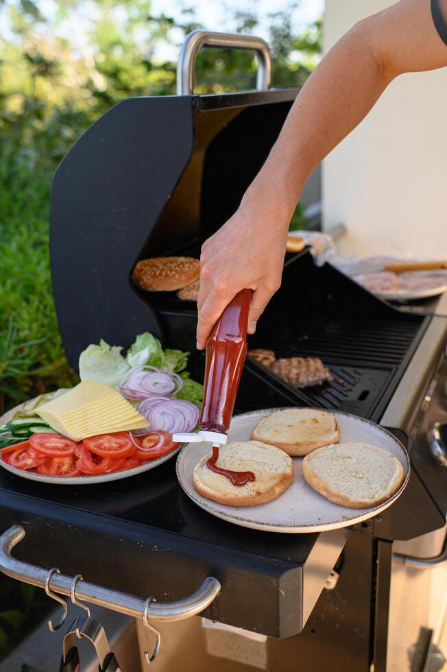 A hand pours ketchup onto toasted burger buns next to a grill where patties are still cooking and a bowl of fresh toppings is ready for assembly. photo