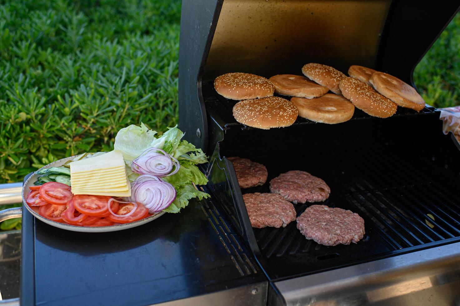 Grill full of sizzling burger patties and toasting sesame buns, with a nearby plate offering crisp lettuce, sliced tomatoes, onions, cheese, and pickles. photo