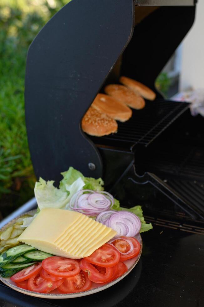 An open grill on a sunny terrace with burger patties and sesame seed buns sizzling, next to a plate of fresh tomatoes, onions, cheese, pickles, and lettuce. photo
