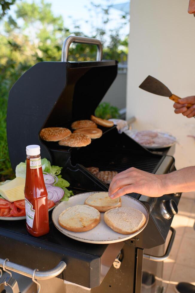 By a busy outdoor grill, hands stack juicy patties and fresh vegetables into burgers, working beside a bowl filled with colorful toppings and cheese. photo