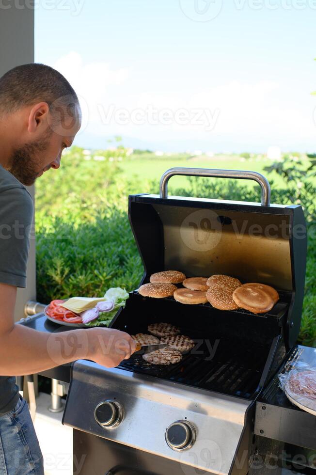 A man tends to burger patties and buns on a sizzling outdoor grill, while a colorful plate of lettuce, tomatoes, onions, pickles, and cheese waits nearby. photo