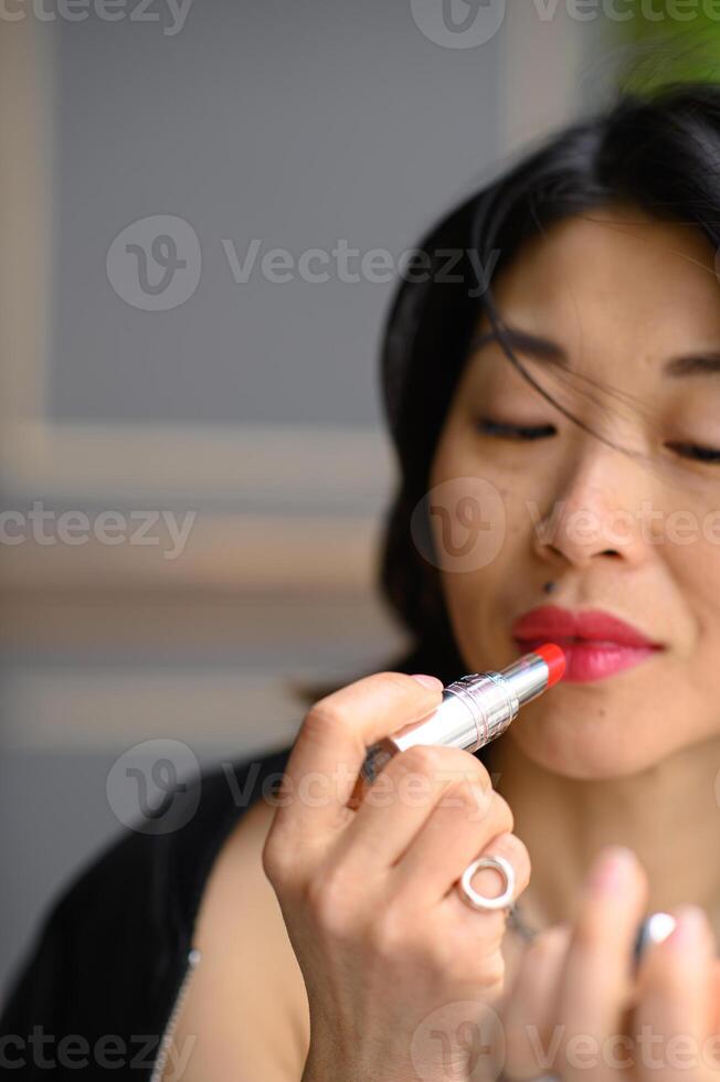 A close-up of an Asian girl as she applies bright red lipstick, her concentration clear in her eyes. The vivid color enhances her natural beauty. photo