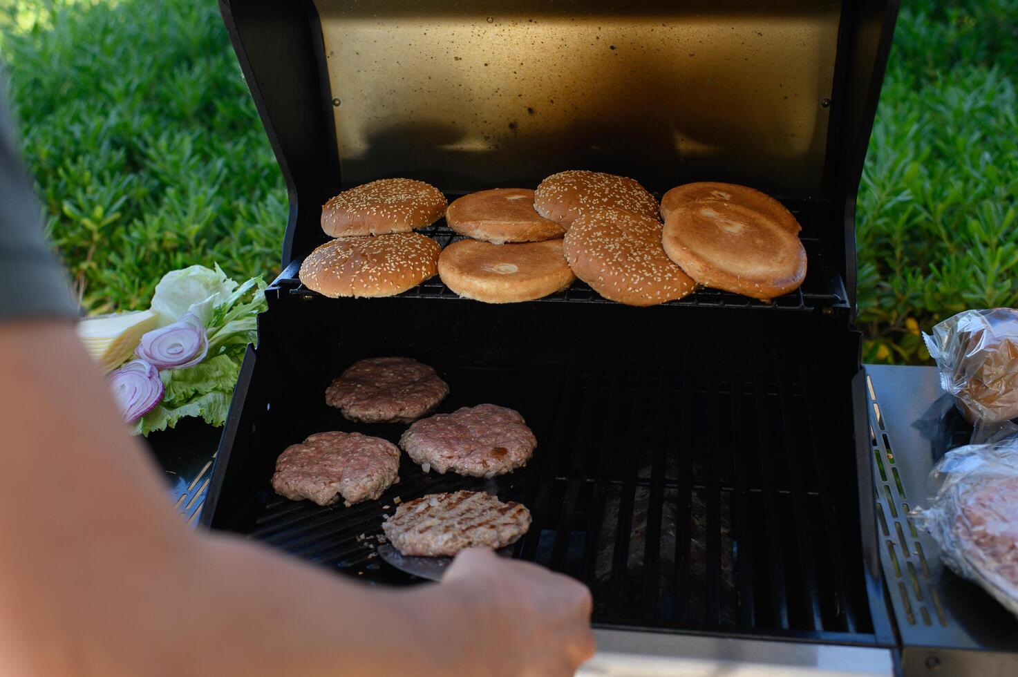 Close-up of hands grilling juicy burger patties over an open flame, with toasted buns and fresh toppings prepared nearby for perfect homemade burgers. photo