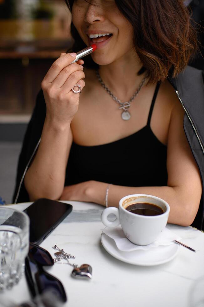 With a calm gaze, a beautiful girl applies red lipstick at a cafe table, her hand resting near a steaming coffee cup. photo