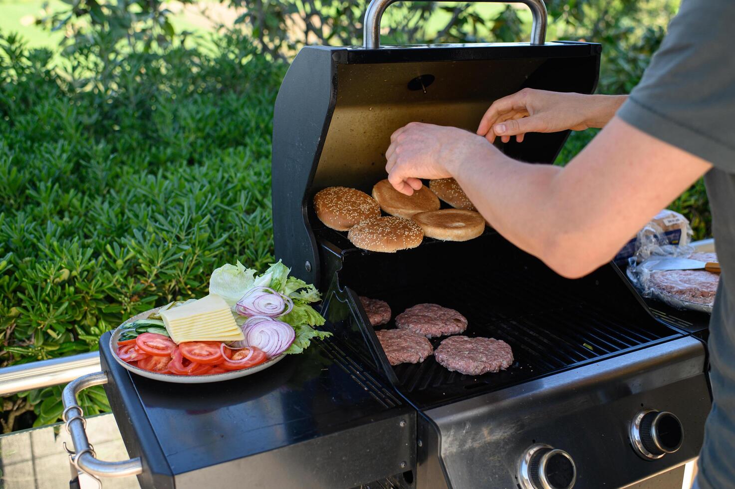Close-up of hands arranging sesame buns on the grill while beef patties cook nearby, and colorful fresh toppings sit prepared on a plate. photo