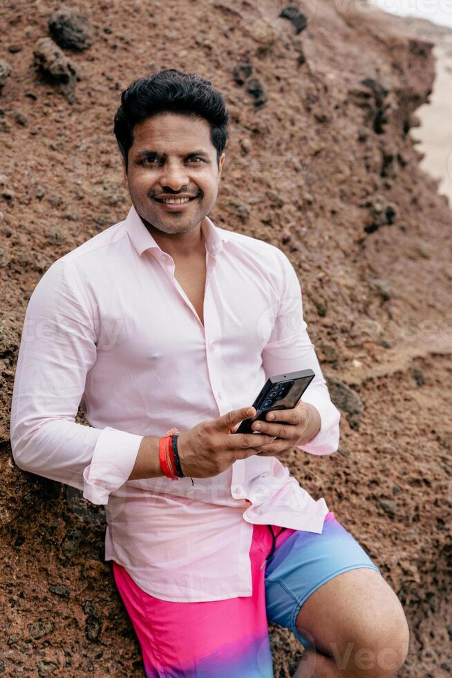 Vertical photo of an Indian businessman at the beach using his smartphone while on vacation