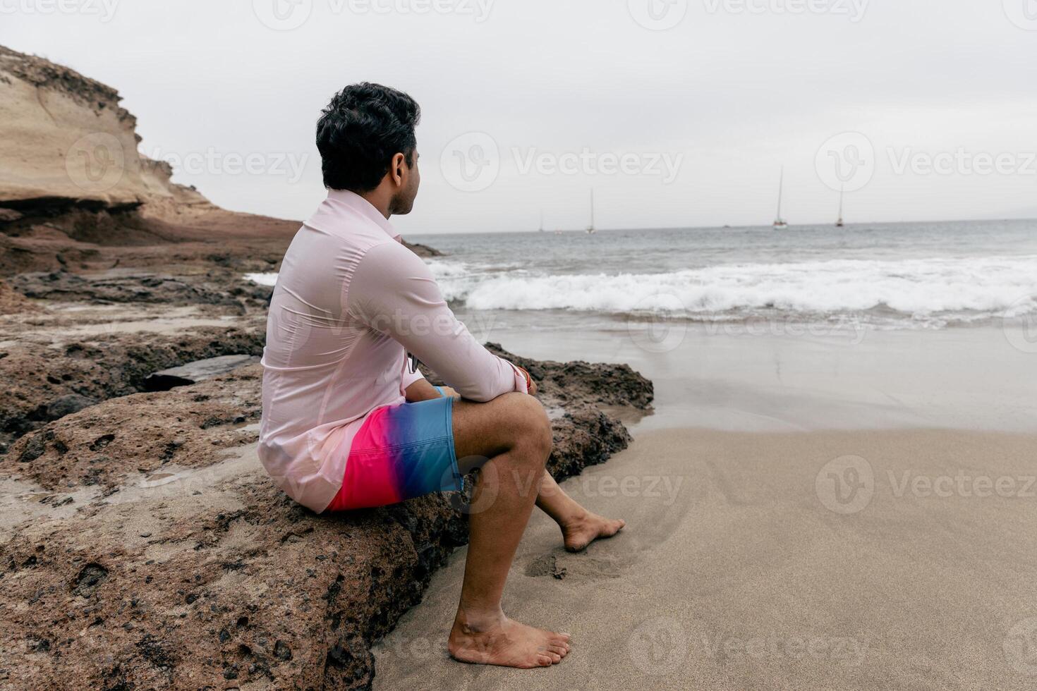 A man sitting on a rock at the beach looking at the ocean and relaxing photo