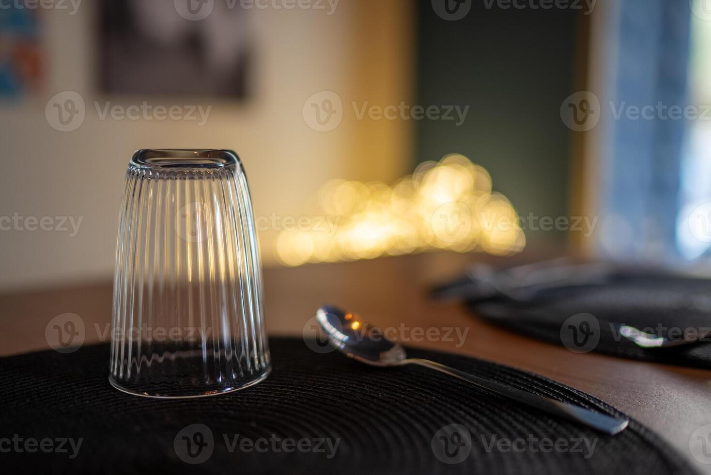 Empty ribbed glass and spoon resting on black placemat on wooden table, blurred background with warm lights and window, elegant and minimalist table setting photo
