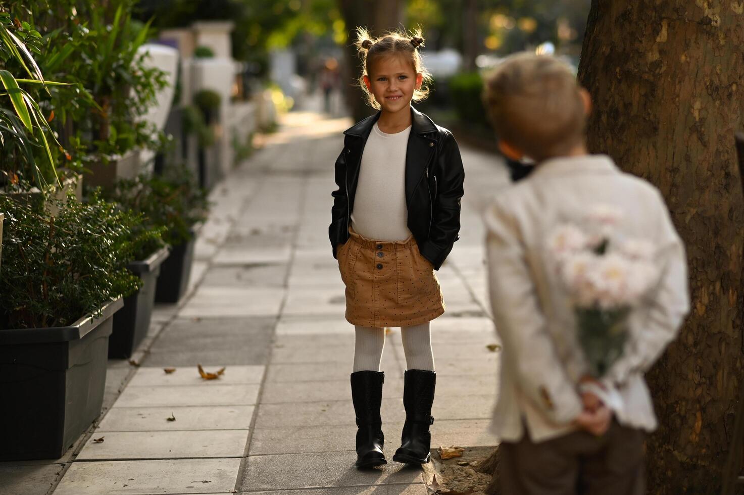 A young boy hides a small bouquet of flowers behind his back while standing in front of a girl at an outdoor cafe photo
