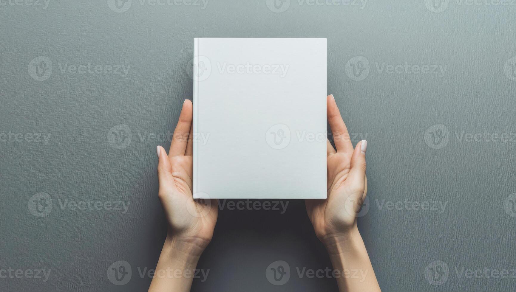 Female hands holding a blank book cover against a gray background, captured from a top view perspective, creating a minimalist mockup photo