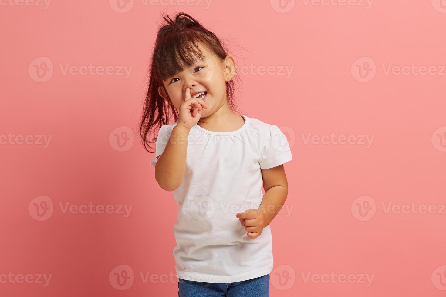 Portrait of little joyful Asian girl picking her nose on pink isolated background. Capricious and stubborn child does not obey his parents. Concept of child crisis of three years. photo