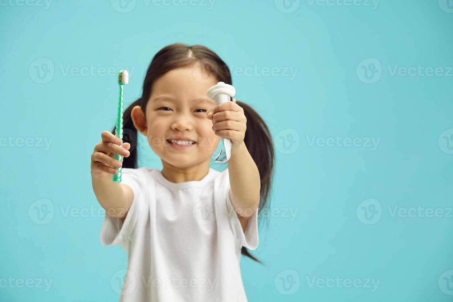 Cheerful and pretty five-year-old girl holding tube of squeezed paste and toothbrush in hands, smiles happily, prepares to brush her teeth, has a beautiful snow-white smile, stands on a blue isolated photo