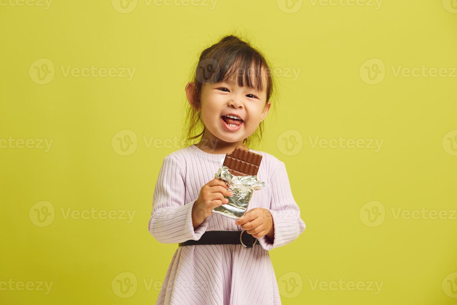 Joyful three year old girl cheerfully and with pleasure eats bar of delicious sweet dark chocolate and smiles funny standing on yellow isolated. photo