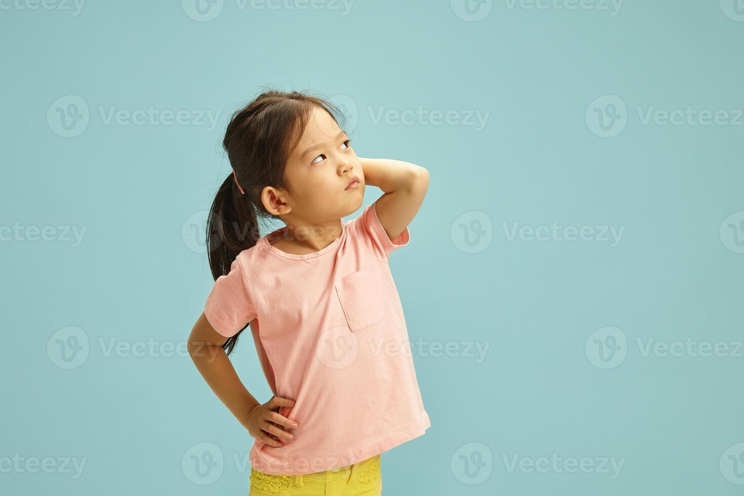 Thoughtful Asian Ethnicity Child Girl Looking Upwards - Aside, Puzzled Scratching Herself Ear, Looks to Empty Copy Space, Standing on Blue Isolated Background photo