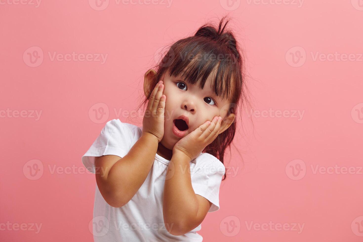 Amazed horrified little three years old girl keeps mouth opened, hands on cheeks, being shocked stares at camera, isolated over pink background. Kid expresses great surprisement. photo
