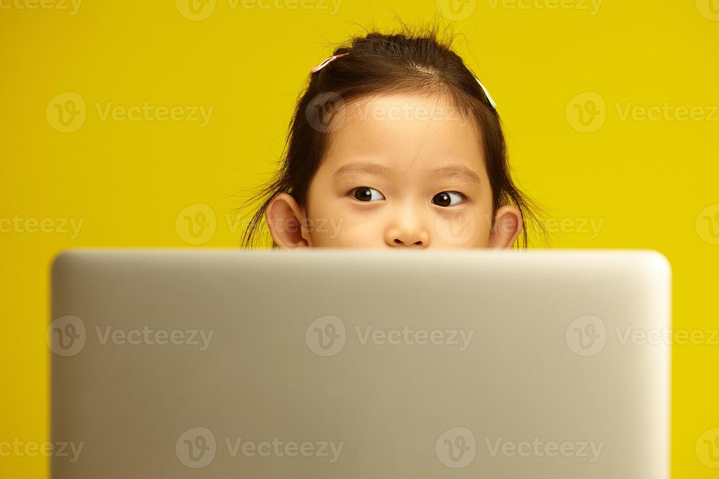 Pensive Asian Preschool Girl with a big opened eyes Engaged in Distance Learning via Laptop sitting against yellow isolated background photo