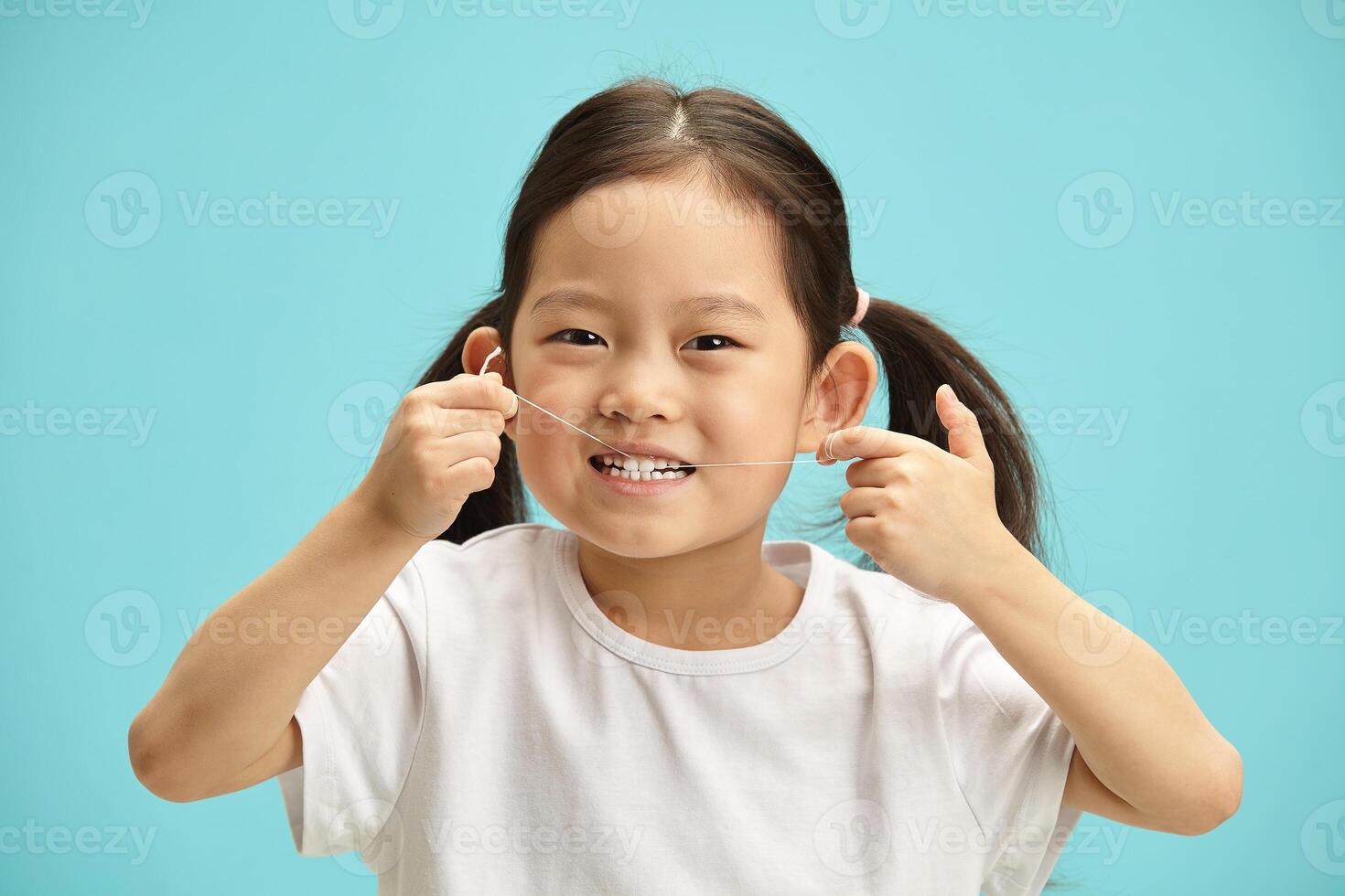 Positive looking little japanese girl does oral care hygiene, cleans her teeth gets rid of food leftovers using dental floss, having satisfied expression, standing over blue isolated background. photo