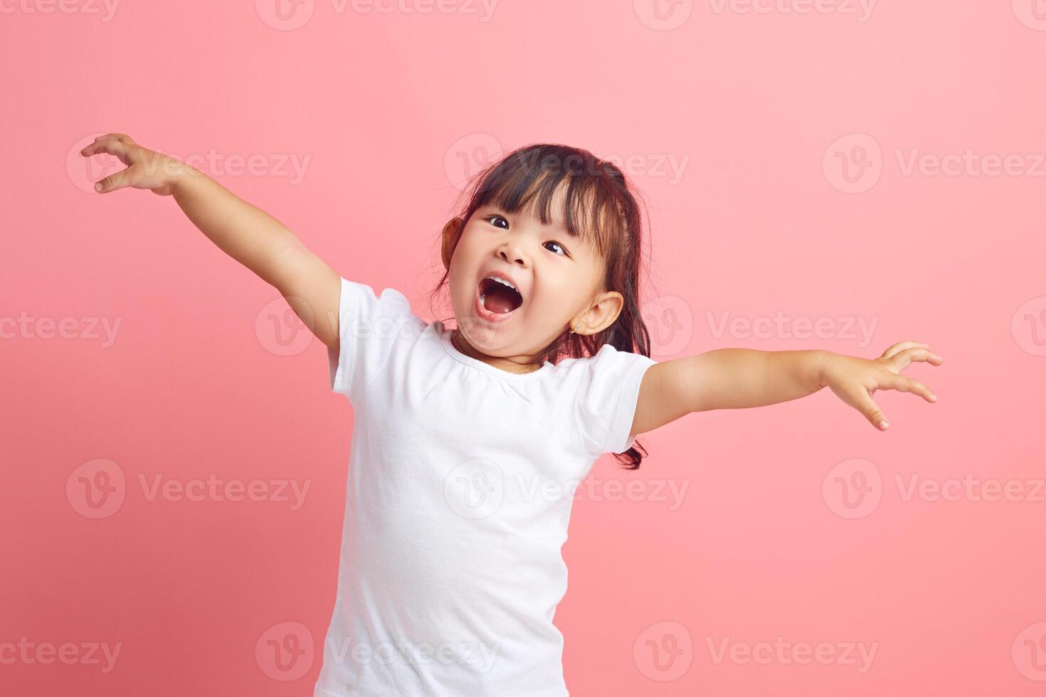 Joyful little Asian girl with her arms outstretched expresses fun and pleasure on isolated pink background. Happy child concept. photo