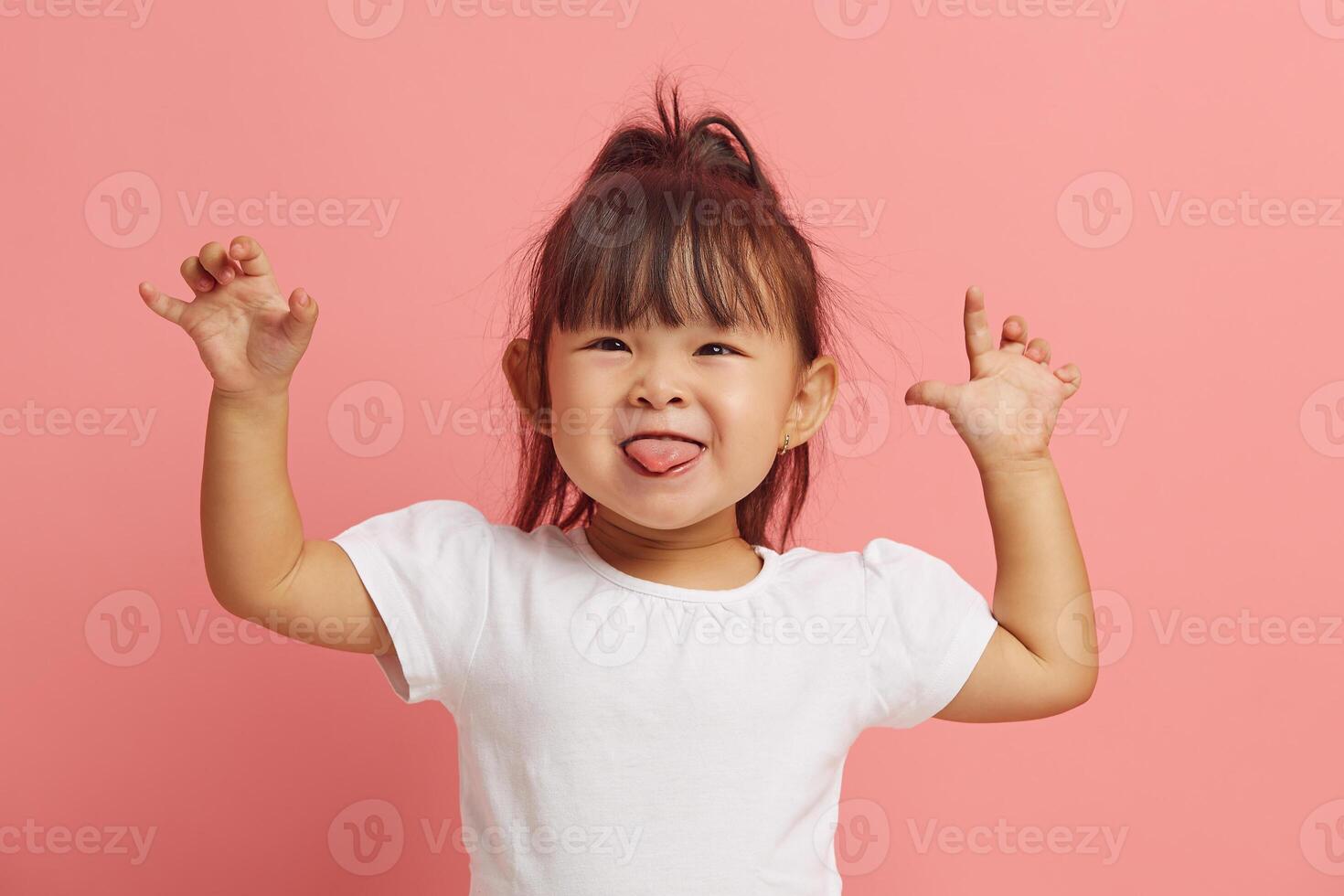 Beautiful little Asian girl has fun and shows her tongue by raising her hands up while standing on a pink isolated. photo