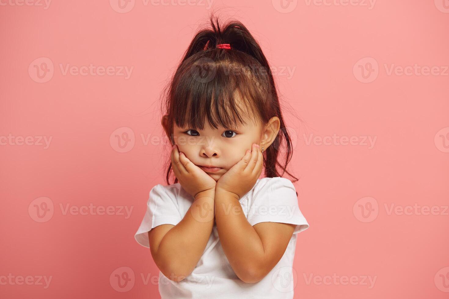 Stunned little three years old asian girl keeps hands on cheeks from wonder standing in white t shirt isolated over pink background. photo